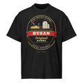 Busan Emblem Tee, front: a gold-and-red Busan crest with Haeundae towers and Gwangan Bridge on a black oversized tee